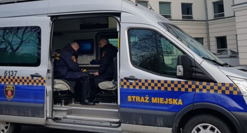 Radiowóz Straży Miejskiej, a w nim funkcjonariusze. W tle budynek.